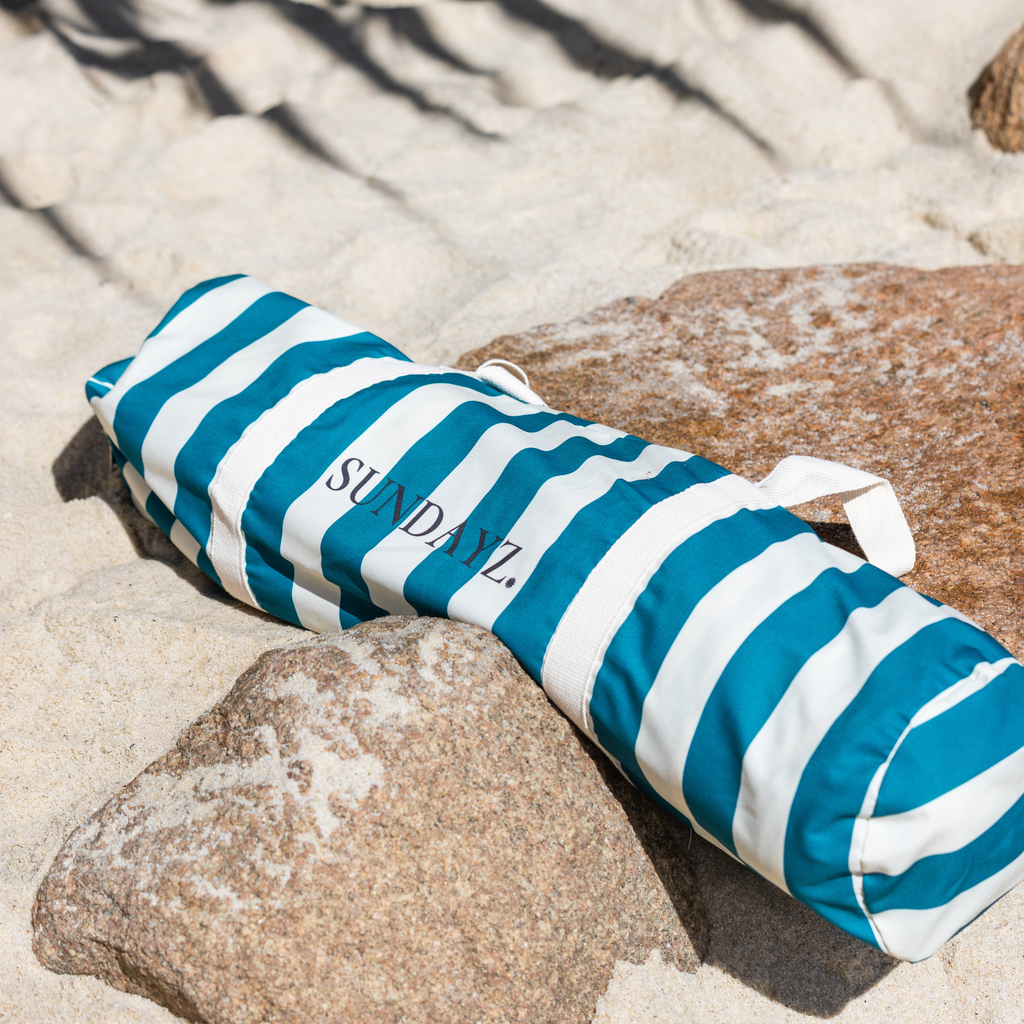 A beach cabana in bag with teal and white stripes lies on sandy ground among rocks. The towel has the brand name 'SUNDAYZ'.
