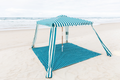 A striped beach cabana with a matching striped beach mat set up on a sandy beach with the ocean in the background.