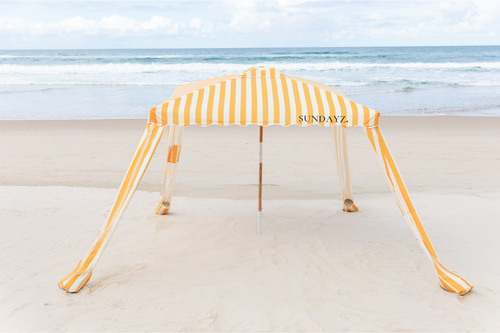 A yellow and white striped beach cabana tent with the brand name 'SUNDAYZ' printed on the top, set up on a sandy beach with the ocean waves in the background.