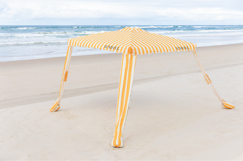 A yellow and white striped beach cabana tent set up on a sandy beach with the ocean and cloudy sky in the background.