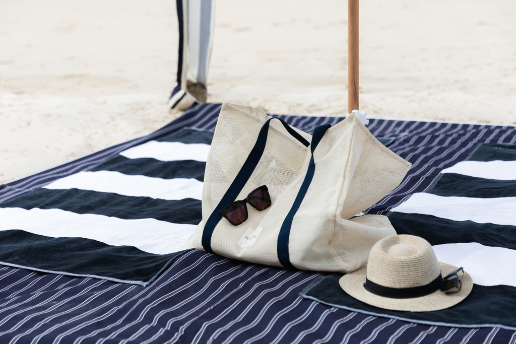 A cream canvas tote bag with navy blue straps sits on a navy and white striped beach mat on the sand, accompanied by a straw hat and sunglasses.